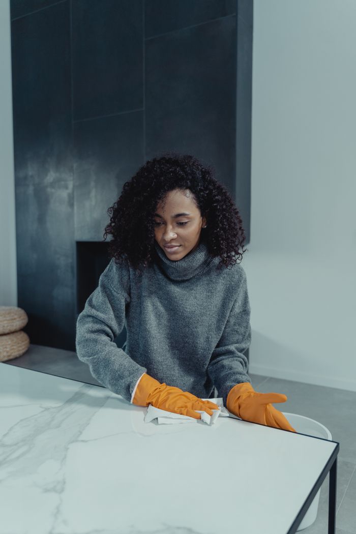 gallery-3 A woman wearing gloves cleans a marble table in a modern indoor setting, focusing on cleanliness and hygiene.
