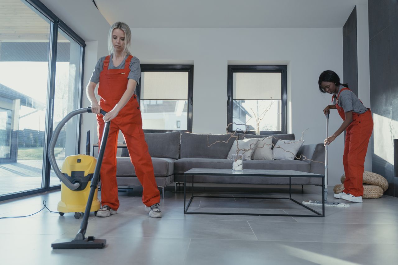 gallery-1 Two women in red overalls vacuuming and sweeping a modern living room.