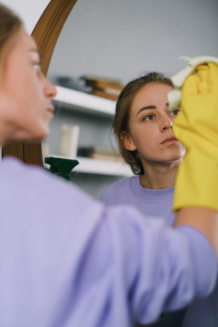services-03 Crop female in purple sweatshirt and latex gloves washing mirror in light apartment in daytime